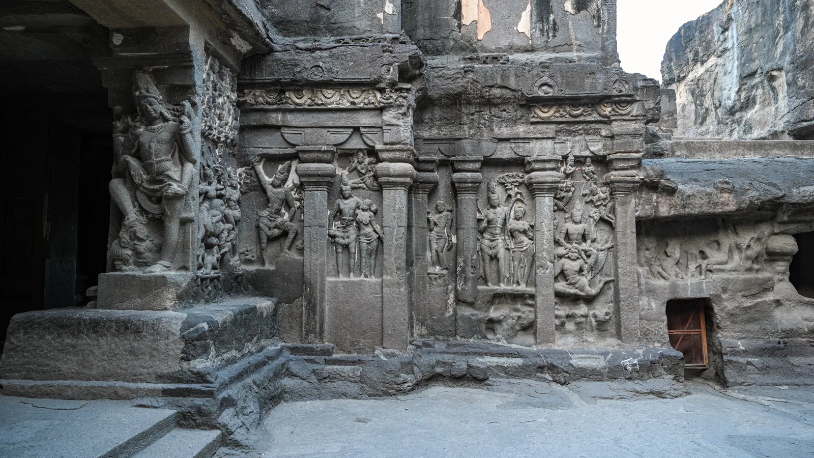 Intricate deity carvings on the walls of the Kailasa Temple — panels of gods, celestial beings, and mythological scenes carved directly into the basalt. Photograph.