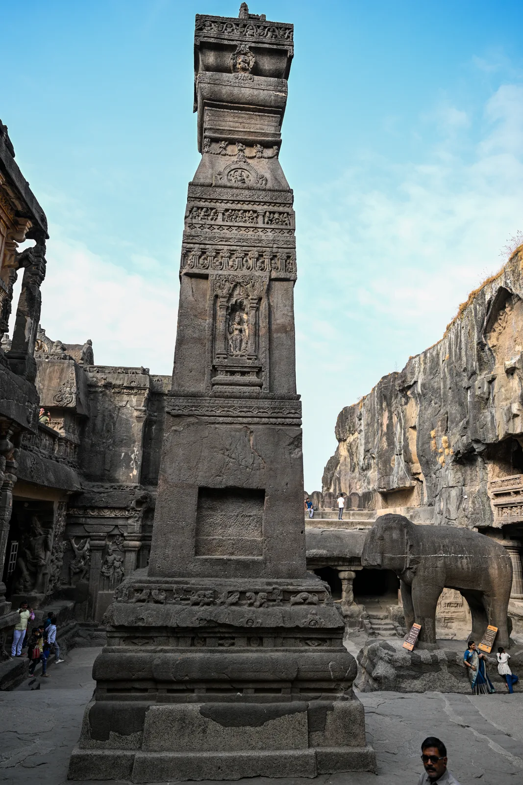 A massive carved pillar with elephant base inside the Kailasa Temple courtyard, with the cliff walls towering above and visitors showing scale. Photograph.